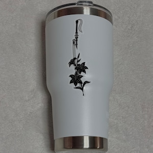 Könnte beinhalten: Weißer isolierter Becher mit einer schwarzen Grafik von Schwert und Blumen. Der Becher hat einen silbernen Rand und Boden sowie einen schwarzen Deckel. Das Design ist eine stilisierte Illustration eines Schwertes und Lilien.