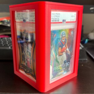 Könnte beinhalten: Ein roter, würfelförmiger Display-Koffer mit zwei bewerteten Sport-Sammelkarten. Die Karten zeigen Footballspieler und sind in klarem Kunststoff eingeschlossen. Der Koffer besteht aus einem soliden, undurchsichtigen roten Material.