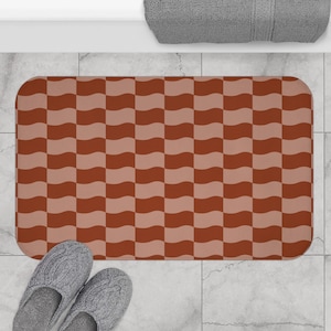 Puede incluir: Una alfombra de baño rectangular con un patrón de tablero de ajedrez ondulado en tonos marrón y beige. La alfombra está colocada sobre un suelo embaldosado, con un par de zapatillas grises visibles en la esquina inferior izquierda. Una toalla gris está en el fondo.