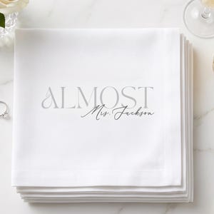 May include: A stack of white fabric napkins featuring the grey printed text ALMOST Mrs. Jackson. The napkins are square with a clean hemmed edge, presented as a set of folded linens for table settings or event decor.