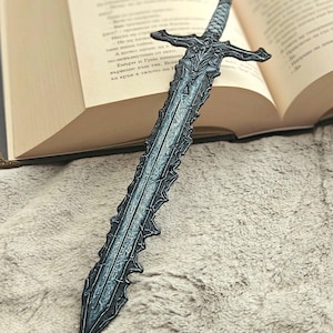 May include: A detailed, dark gray sword-shaped bookmark with intricate designs. The bookmark is placed in an open book with text visible on the pages. The sword has a cross-shaped hilt and a textured blade, adding a fantasy element.