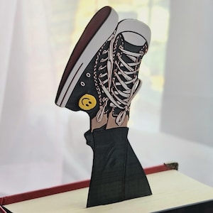 May include: A flat, vertical bookmark shaped like a black high-top sneaker with white laces, a white toe cap, and a yellow smiley face icon on the side. The sneaker is attached to a black, triangular base that inserts into the pages of a book.
