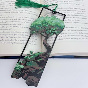 May include: A black metal bookmark with a tree design. The tree has green leaves and a brown trunk, with a green tassel at the top. The bookmark is placed in an open book.