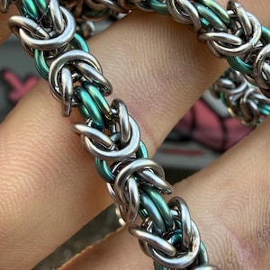 May include: Close-up of a chainmail bracelet with alternating links of silver and teal. The intricate design features a byzantine weave, creating a textured and eye-catching accessory. The bracelet is made of metal.