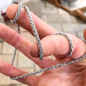 May include: A silver chain necklace with a woven design. The chain is shown wrapped around a person's fingers.