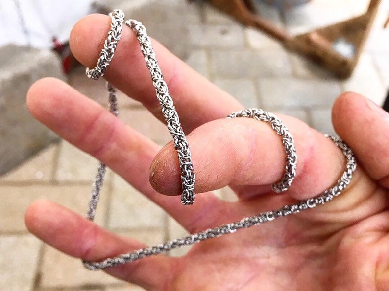 May include: A silver chain necklace with a woven design. The chain is shown wrapped around a person's fingers.