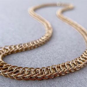 Peut inclure: Un collier en chaîne dorée composé d'anneaux entrelacés. La chaîne est posée sur une surface grise.