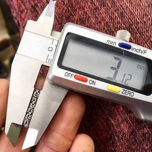 May include: A silver digital caliper measuring a silver chain link bracelet. The caliper reads 3.12 mm.