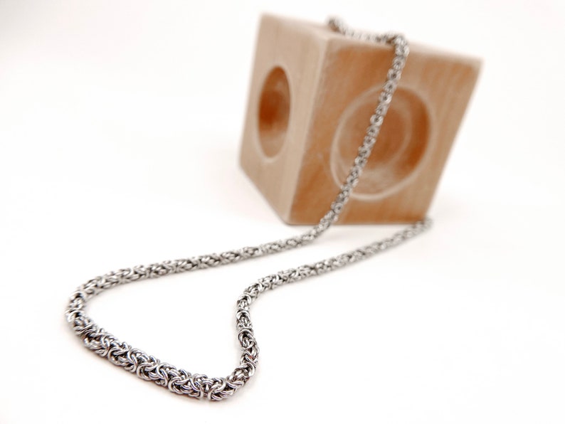 May include: A silver chain necklace with a woven design. The chain is draped over a wooden block.