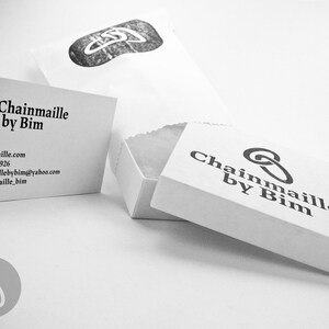 May include: A black and white business card with the text "Chainmaille by Bim" and contact information. The card is next to a white box with the same text on it.