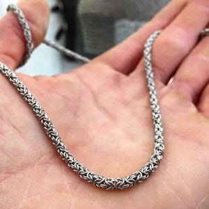 May include: A silver chain necklace with a woven design. The chain is made of small, interlocking links.