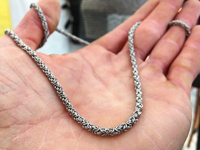 May include: A silver chain necklace with a woven design. The chain is made of small, interlocking links.