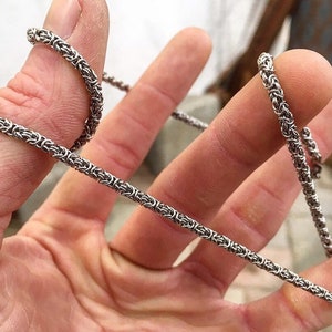 May include: A silver chain necklace with a woven design. The chain is shown wrapped around a finger.