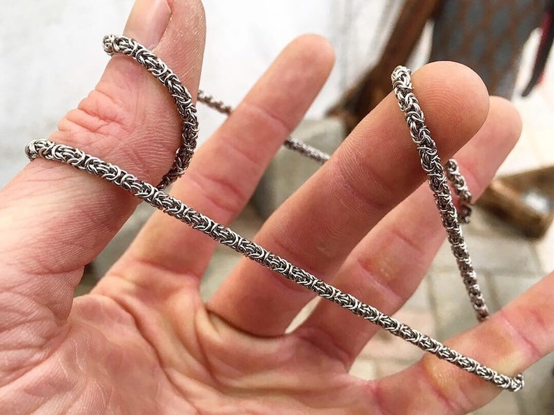 May include: A silver chain necklace with a woven design. The chain is shown wrapped around a finger.