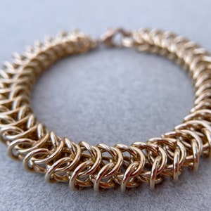 Peut inclure: Un bracelet en chaîne dorée avec un motif tissé. Le bracelet est composé de petits maillons métalliques interconnectés.