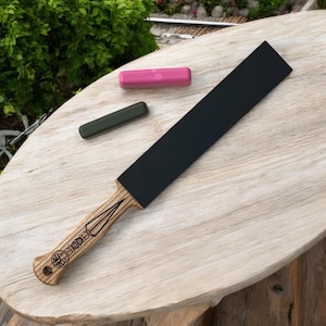 May include: A black leather strop with a wooden handle. The handle features a decorative spearhead design. The strop is on a light wood surface with a pink and green leather strop alongside it.