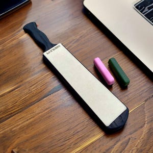 Puede incluir: Un acero de afilar negro y gris con un mango negro y la palabra "Premium" impresa en el lateral. Dos pequeñas piedras de afilar de colores, una rosa y una verde, están al lado del acero.