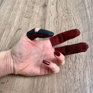 Puede incluir: Tres protectores de dedos individuales hechos de tela color granate oscuro. El protector para el pulgar presenta una punta con textura acanalada negra, mientras que los protectores para el índice y el medio son completamente granates.