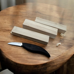 Puede incluir: Tres bloques de madera de color marrón claro y un pequeño cuchillo con mango negro con la palabra "Pferd" en la hoja. Los bloques están dispuestos en forma triangular sobre una superficie de madera.