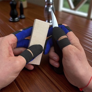 May include: A person wearing blue and black finger protection gloves is sharpening a knife with a whetstone. The gloves have a blue and black design with finger openings for dexterity. The knife has a silver blade.