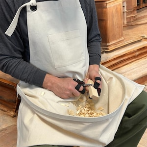 May include: A white canvas apron with a large pocket and a gathered bottom. The apron is worn by a person who is using a knife to carve a piece of wood. Wood shavings are scattered around the person's feet.
