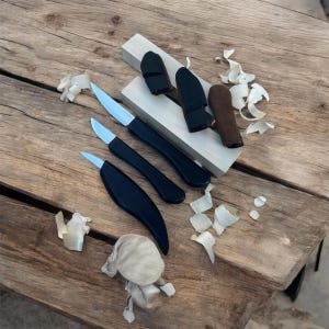 May include: Three small carving knives with black handles and silver blades are displayed on a wooden surface. The knives are in leather sheaths.