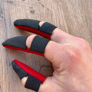 May include: Three fingertip protectors with black fabric and red padding. The protectors are worn on the index, middle, and ring fingers.