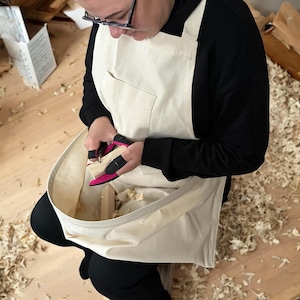 May include: A person wearing a white apron with a large pocket and black sleeves is working on a piece of wood with pink finger guards. The apron has a large pouch in the front to catch wood shavings.