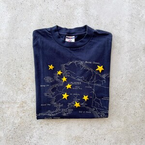 May include: Navy blue t-shirt featuring a map of Alaska with yellow star accents. The map includes geographical details and the text "Alaska". A classic crew neck tee, perfect for casual wear.