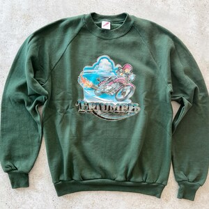 K&ouml;nnte beinhalten: Ein dunkelgr&uuml;ner Pullover mit einem Aufdruck eines Comic-Charakters, der ein Motorrad f&auml;hrt, mit dem Wort "Triumph" auf der Vorderseite.