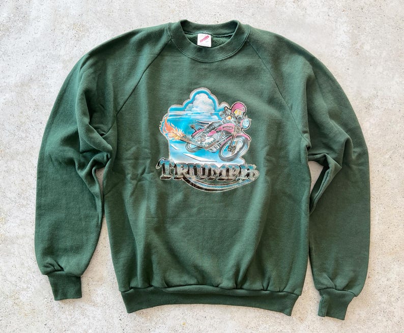 K&ouml;nnte beinhalten: Ein dunkelgr&uuml;ner Pullover mit einem Aufdruck eines Comic-Charakters, der ein Motorrad f&auml;hrt, mit dem Wort "Triumph" auf der Vorderseite.