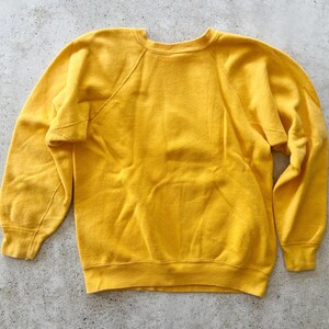 May include: A solid mustard yellow long-sleeve sweatshirt featuring a classic crew neckline, raglan sleeves, and ribbed cuffs at the wrists and hem. The garment is constructed from a soft, fleece-like material with a plain, unpatterned design.