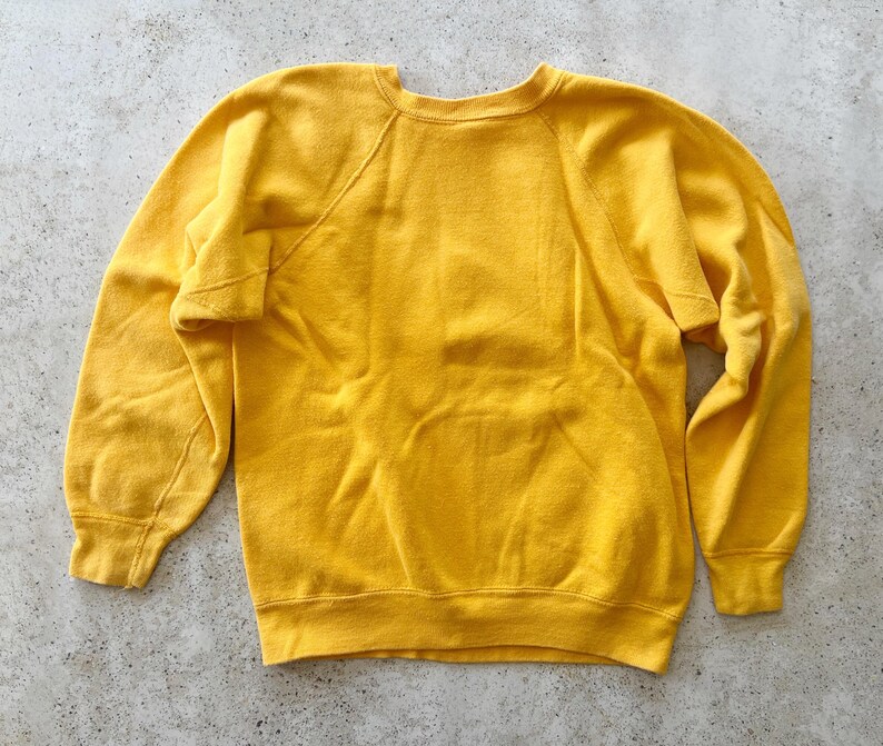 May include: A solid mustard yellow long-sleeve sweatshirt featuring a classic crew neckline, raglan sleeves, and ribbed cuffs at the wrists and hem. The garment is constructed from a soft, fleece-like material with a plain, unpatterned design.