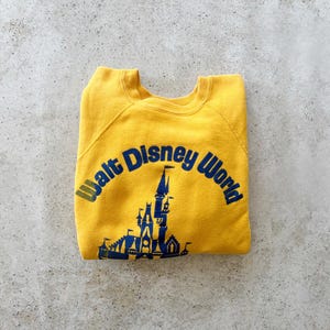 May include: A mustard yellow crewneck sweatshirt featuring a dark blue graphic print of the Walt Disney World castle and the text Walt Disney World arched above the castle. The garment has long sleeves and a ribbed collar, cuffs, and hem.