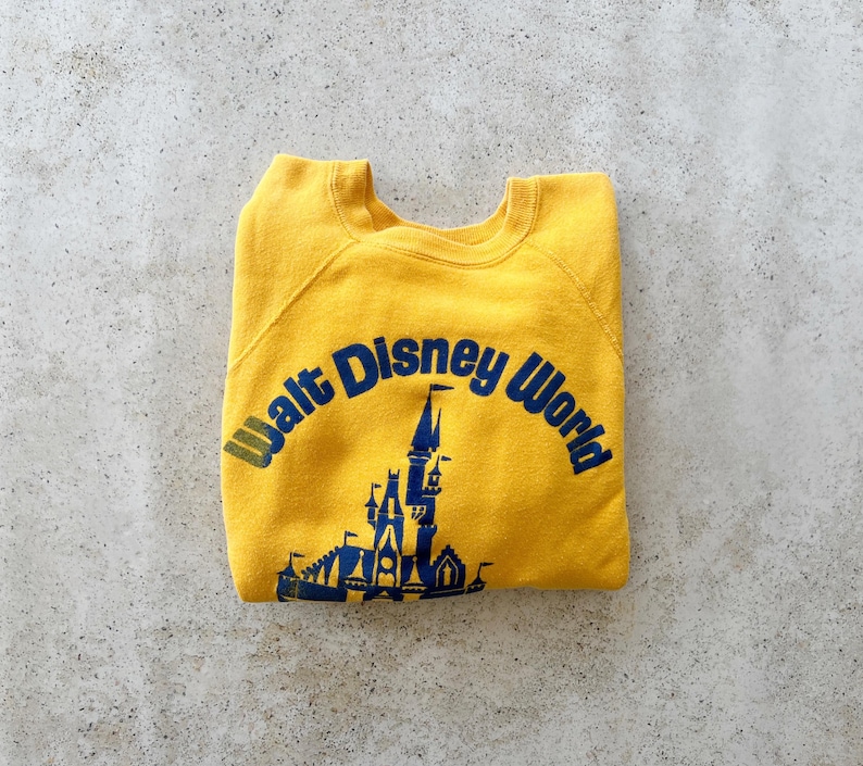 May include: A mustard yellow crewneck sweatshirt featuring a dark blue graphic print of the Walt Disney World castle and the text Walt Disney World arched above the castle. The garment has long sleeves and a ribbed collar, cuffs, and hem.