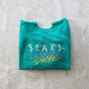 May include: Teal sweatshirt with white embroidered figures and the text "LAMBDA Delta" in pink and yellow. The sweatshirt is folded and lies on a gray surface. The figures appear to be dancing or in motion.
