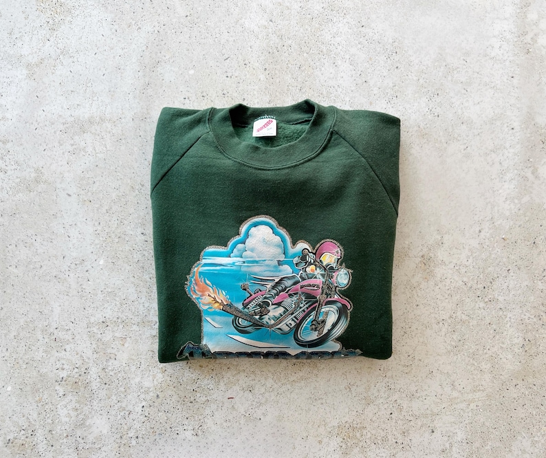 K&ouml;nnte beinhalten: Ein dunkelgr&uuml;ner Sweatshirt mit einem Aufdruck einer Person, die ein Motorrad f&auml;hrt, vor einem blauen Himmel mit Wolken.