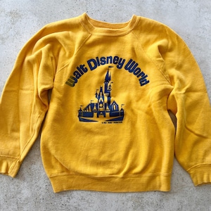 May include: A bright yellow long-sleeve sweatshirt featuring a blue graphic print of the Walt Disney World castle logo on the front. The design includes the text Walt Disney World arched over the castle illustration with smaller text below.
