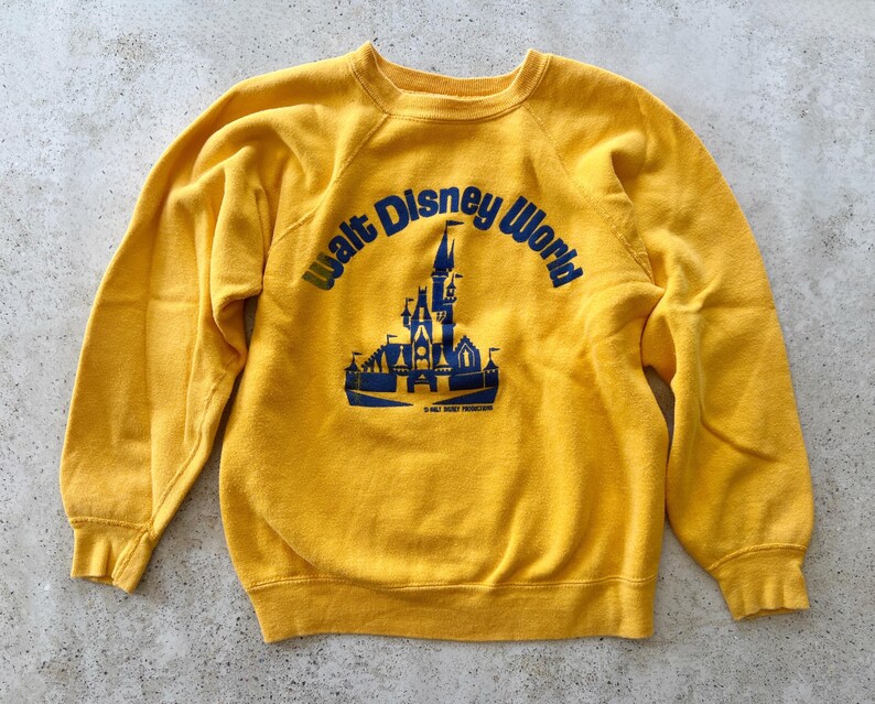 May include: A bright yellow long-sleeve sweatshirt featuring a blue graphic print of the Walt Disney World castle logo on the front. The design includes the text Walt Disney World arched over the castle illustration with smaller text below.