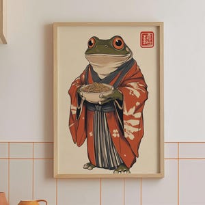 Może przedstawiać: Oprawiony wydruk przedstawiający kreskówkową żabę w czerwonym kimono trzymającą miskę z makaronem. Żaba ma duże czerwone oczy i jest umieszczona na kremowym tle. Czerwona pieczęć znajduje się w prawym górnym rogu.