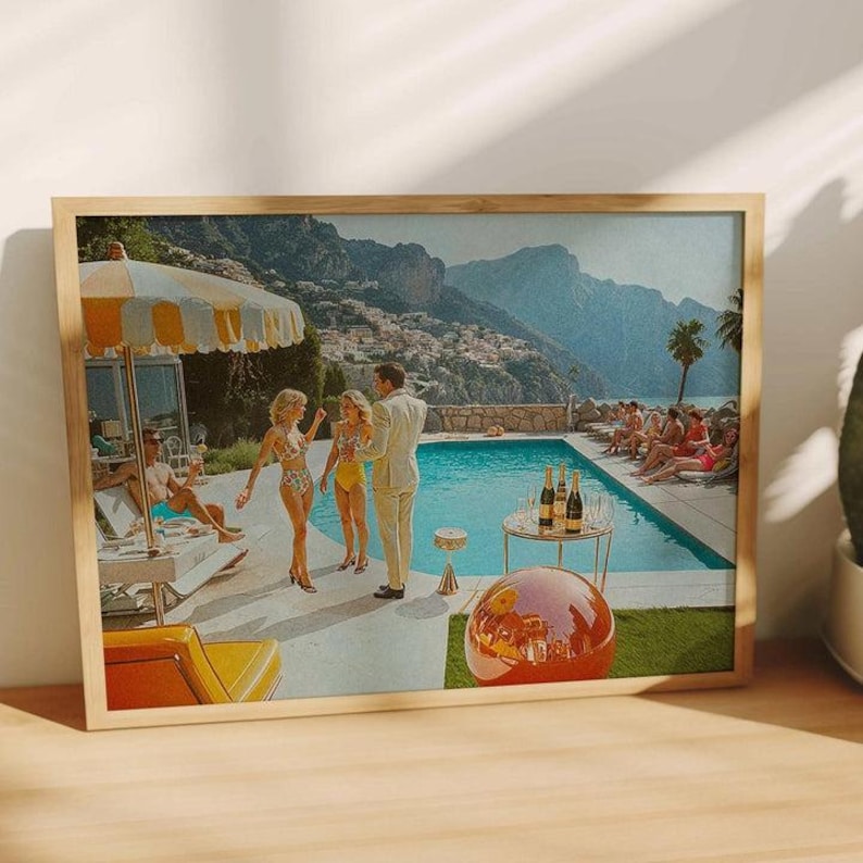 May include: A framed print depicting a retro pool party scene. The image features people in swimwear, a pool, and a mountain backdrop. A table with bottles and glasses is in the foreground, with a large orange sphere. The print is in a wooden frame.