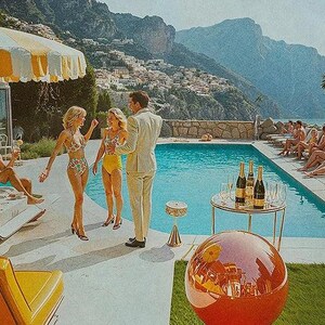 May include: Vintage-style image of a poolside gathering. People in swimwear and suits are socializing near a pool with champagne bottles on a side table. The scene is set against a backdrop of mountains and a coastal town.
