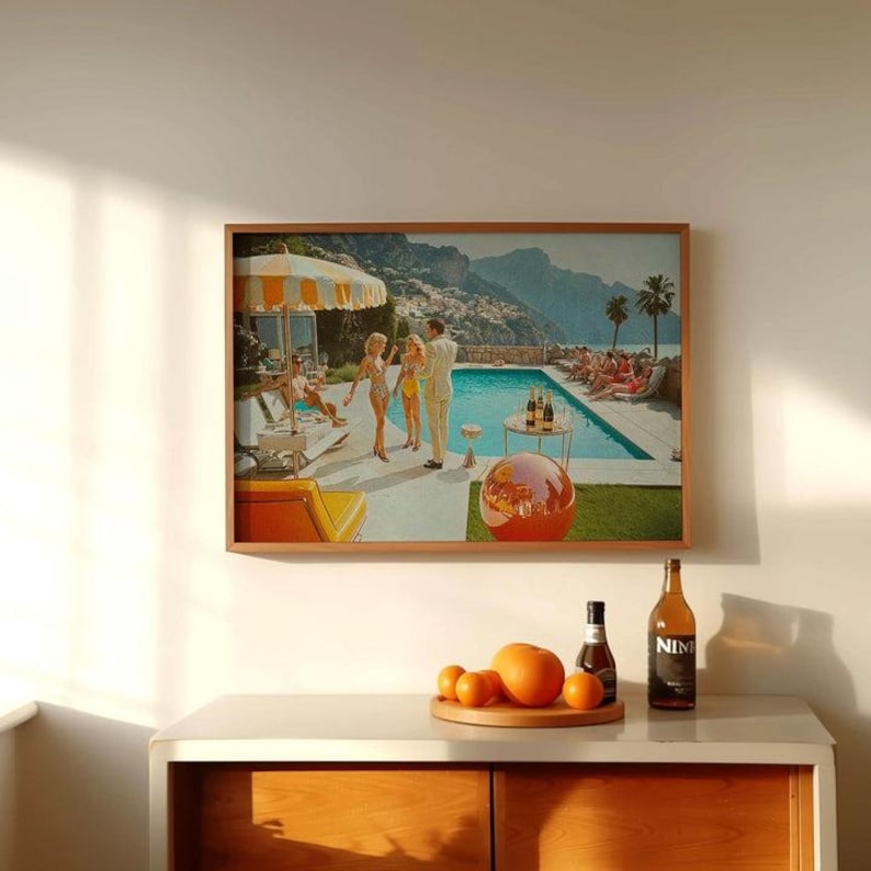 May include: A framed print depicting a retro pool party scene with people in swimwear, a pool, and a mountain backdrop. The print is in a wooden frame. A wooden tray with oranges and a bottle of wine are on a white cabinet.