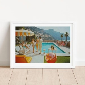 May include: A framed print depicting a vintage pool party scene. The artwork features a swimming pool, people in swimwear, a striped umbrella, and a red and orange sphere. Palm trees and mountains are visible in the background.