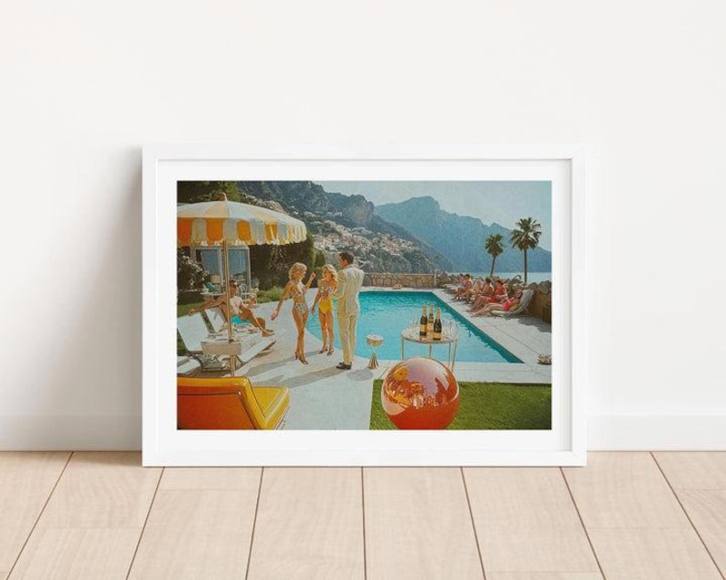 May include: A framed print depicting a vintage pool party scene. The artwork features a swimming pool, people in swimwear, a striped umbrella, and a red and orange sphere. Palm trees and mountains are visible in the background.