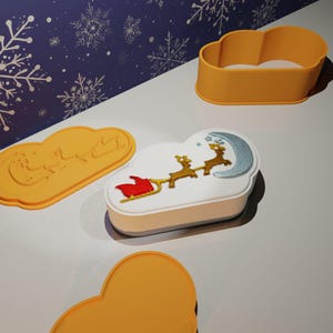May include: A festive Christmas-themed cookie with a cloud shape, decorated with a sleigh, reindeer, and a crescent moon. The cookie is next to cloud-shaped cookie cutters in yellow. The background is a blue surface with white snowflake patterns.