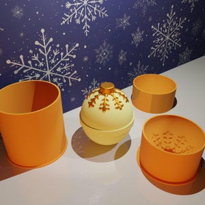 Könnte beinhalten: Eine weiße und goldene Weihnachtskugel-Badebombe mit Schneeflockenmuster. Die Badebombe ist von drei orangefarbenen Silikonformen umgeben.