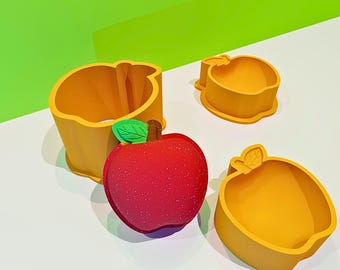 May include: An arrangement of apple-shaped items. A red, textured apple with a green leaf and stem sits next to three yellow apple-shaped molds. The molds are in different sizes and shapes, suggesting they are for baking or crafting.
