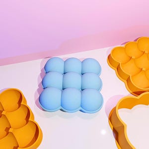 May include: A light blue silicone ice cube tray with nine spherical molds. Two orange silicone molds are also visible. The items are on a white surface with a pink and purple background.
