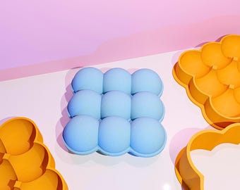 May include: A light blue silicone ice cube tray with nine spherical molds. Two orange silicone molds are also visible. The items are on a white surface with a pink and purple background.